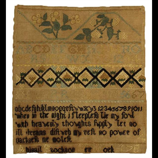 A now-yellowed cloth sampler created by a member of the Dunbar family. Magnolia blossoms are stitched on the top row, followed by a row for the alphabet in capital letters. The third row is a diamond pattern in dark thread around white centers. The fourth row features a lowercase English alphabet, followed by “When in the night I sleepless lie my Lord with heavenly thoughts supply / let no ill dreams disturb my rest / no power of darkness me molest" followed by "Abigail Stockton Her Work / 1784."