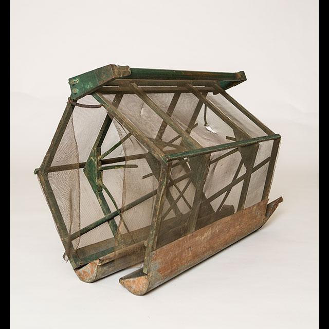 A color photograph of a boll weevil trap. The body of the trap is made of green-painted wood. The frame is covered in metal screening. There are two metal troughs on the base. 