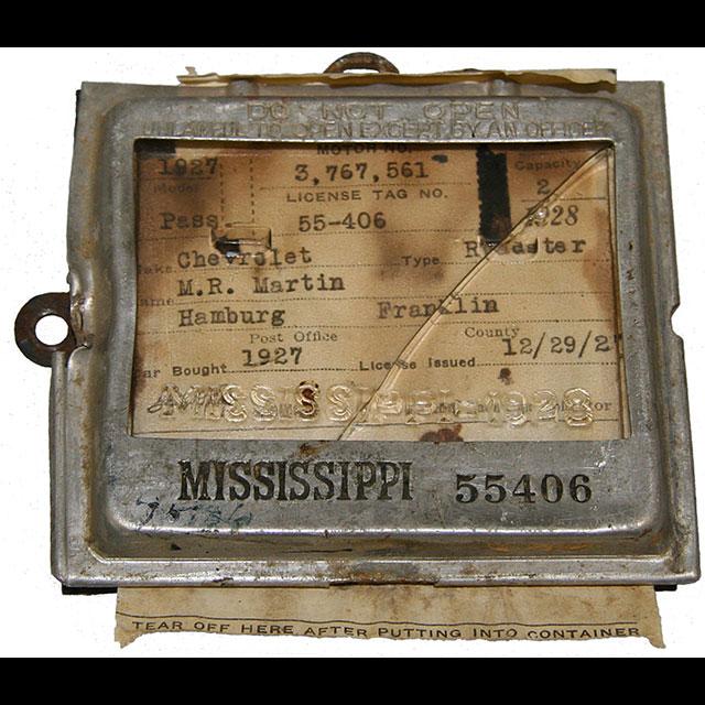 A color photograph of a vehicle title and registration inside a display holder. The holder was meant to be mounted to a vehicle’s dashboard and only be opened by law enforcement. It has a silver metal frame with a glass front. The car’s title is yellowed paper with information about the car and owner printed in black text. 