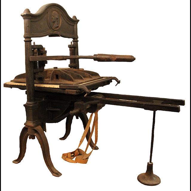 A color photograph of a printing press. The brown, iron press has four legs joined by a curved bar in the center. A bas-relief medallion of the bust of George Washington in profile is pressed across the top bar of the frame. 