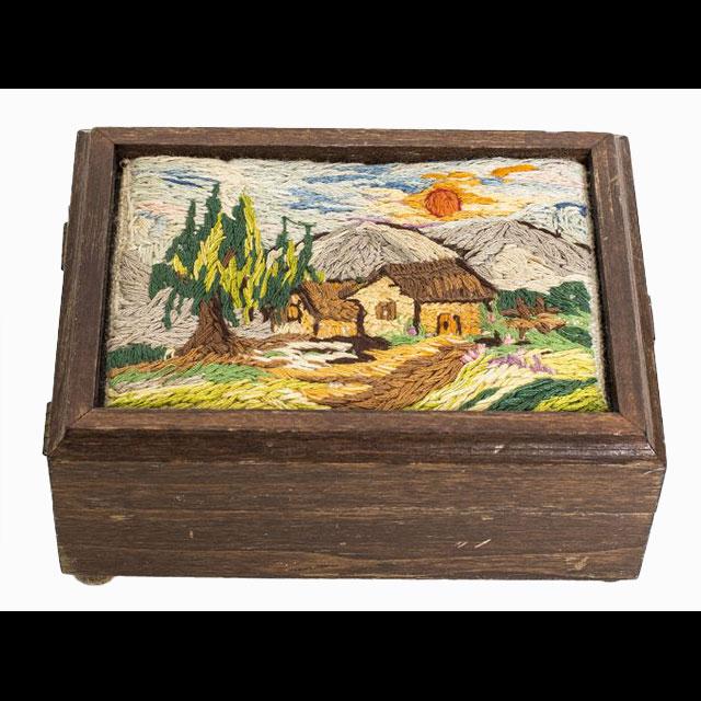 A color photograph of a wood box with Wilda Cartagena’s embroidery. The box is made of dark wood and has a lock on the front. It rests on four round legs. The embroidery on top depicts two houses in front of a rising sun in the mountains.