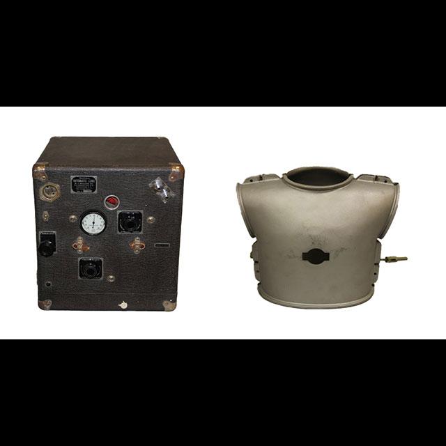 A color photograph of an iron lung portable pump (left) and body plate (right). The pump is encased in a dark box with gold metal corners. There is a white gauge in the center with three black knobs around it. The body plate is in the shape of a hollow child’s torso. It is made of silver metal, and has holes for the patient’s head and arms, and a hole in the center for the pump to attach.