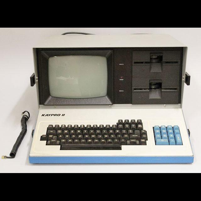 A color photograph of the KAYPRO II computer. The border and number keys are light blue. The main keyboard, drivers, and power cord are black. The main body is white. The unit was made of metal and has a built-in monitor. 