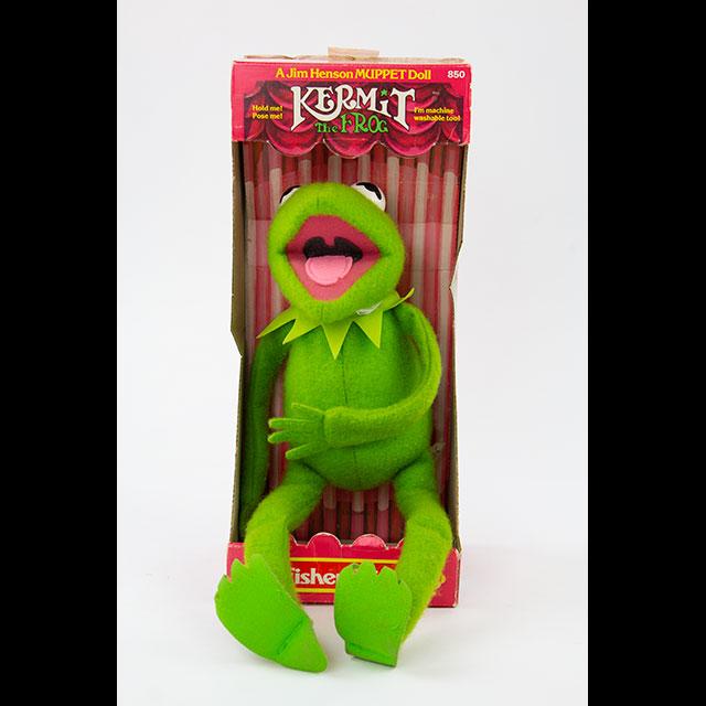 A color photograph of a Kermit the Frog doll. The body, head, arms, and legs are bright green felt. The mouth is open and has red felt for the inside of the mouth, black felt in the back of the mouth, and a light pink felt tongue. The eyes are black and white and are visible at the top of the head. Kermit is housed inside his original box, which is pinkish red and 15.25” tall by 6.5” wide and 10.25” deep. The front of the box is open where Kermit sits.