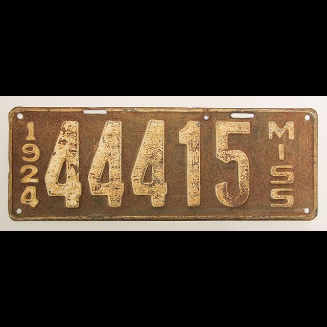 A color photograph of a 1924 license plate. The license plate is metal. The letters and numbers were painted white and the background and border around the edge are rust colored. The left side of the plate shows the year, 1924. The center has the plate number, “44415,” and the right side reads: “MISS.”