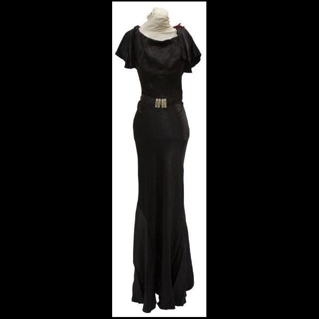 A color photograph of Boyette’s pageant dress. The dress is floor length and made of black satin with fluttery, short sleeves and a scoop neckline. The dress has a matching black belt with a gold buckle. 