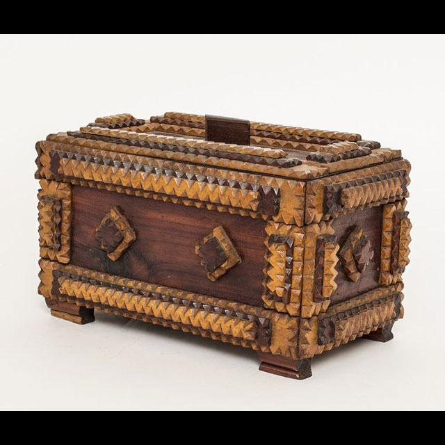 A color photograph of a POW-made trinket box. The box is made of wood in various shades of brown and has four feet. There is an ornate zigzag design on the trim, and the box has a diamond pattern throughout.