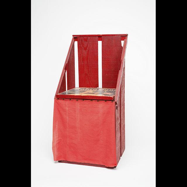 A color photograph of a potty chair. The chair is made using parts from a Robin Hood brand orange crate. It has a back rest and a lid featuring an illustration of Robin Hood covers the seat. The lid is removable and a red curtain below it conceals a compartment for a chamber pot.