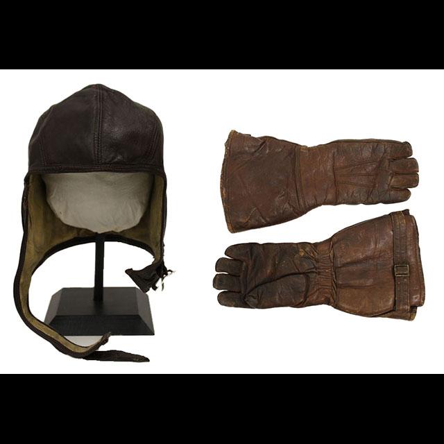 A color photograph of John C. Robinson’s flight helmet and gloves. Both are made from dark brown leather. The cap is lined with cream-colored suede. It was secured with a strap on the left inserted into a metal clasp on the right. The gloves feature metal closures near the wrist.