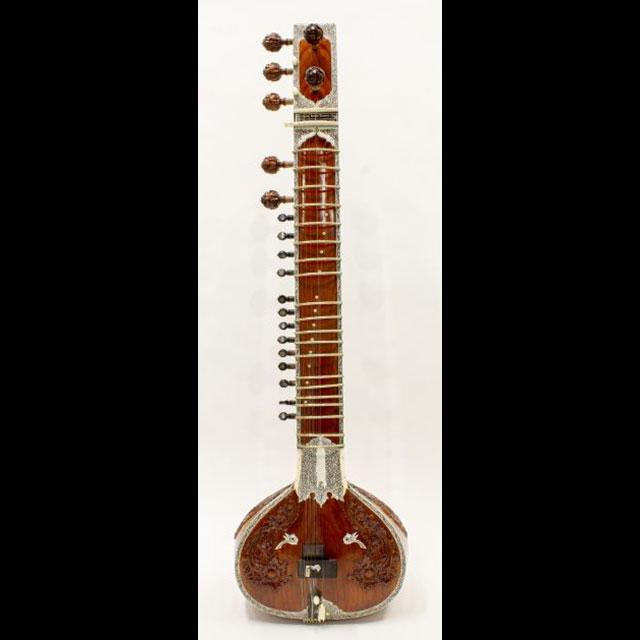 A color photograph of a sitar. The neck of the instrument is varnished wood with detailed carvings and decoration. There are thirteen small tuning pegs on the side and seven larger tuning pegs near the top of the neck. The instrument is strung down the center with gold string. The body is made from a pumpkin that is coated in a brown-orange varnish. 