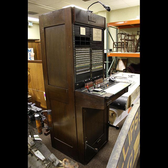 A color photograph of an early 20th-century general store switchboard, positioned at a three-quarter profile. The cabinet is made of dark colored wood. There is a lamp at the top of the cabinet. The top half of the cabinet houses the main frame of the board. The controls are installed in the exterior component in the center.