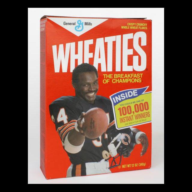A color photograph of a box of Wheaties cereal featuring Walter Payton. The box is orange, with white and yellow text. Payton is in the center of the box wearing his football uniform and pointing a football to the viewer. There is a graphic advertising a contest to the right of him. 