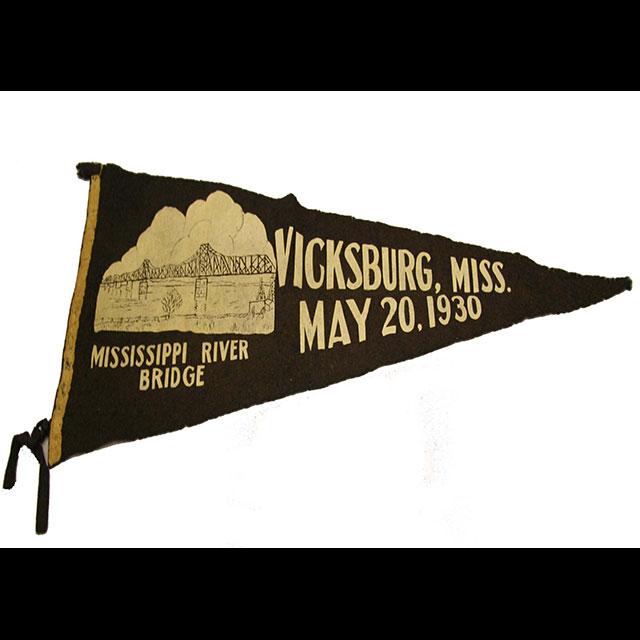 A color photograph of a commemorative pennant celebrating the opening of the Mississippi River Bridge. The pennant is navy blue felt and triangular in shape. An image of the bridge and its opening date were painted in white. There is a white border on the left side and a string to tie the pennant to a pole. 